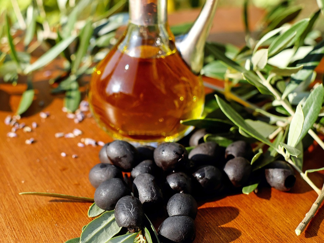 Eine Flasche Olivenöl steht auf einem Holztisch, umringt von frischen Oliven, Meersalz und Kräutern. 